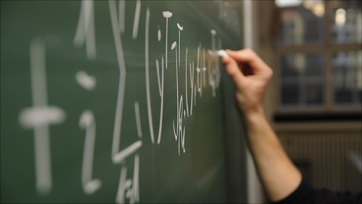 Seitliche Nahaufnahme einer Hand, die etwas mit Kreide an eine Tafel schreibt.