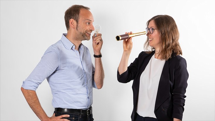 Ein Mann und eine Frau stehen sich grinsend gegenüber. Der Mann hält eine Glühbirne vor sein Gesicht. Die Frau hält ein altmodisches Fernglas vor ihr Auge.