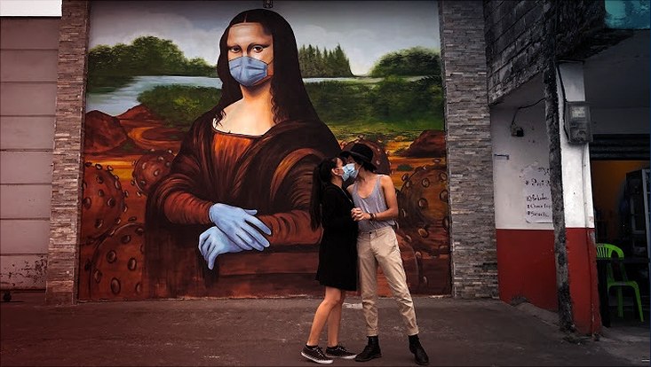 Ein sich mit medizinischer Maske küssendes Paar steht vor Streetart (Mona-Lisa mit Mund-Nasen-Schutz und Handschuhen)
