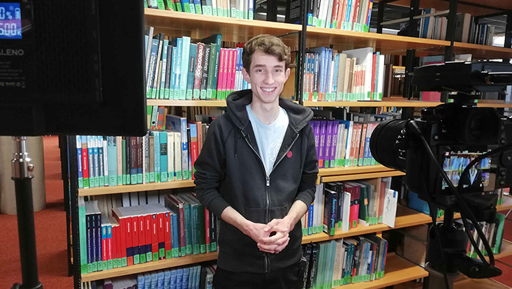 Studierender steht vor einem Bücherregal. Vor ihm ist ein Kamera-Equipment aufgestellt.
