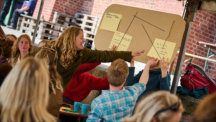 Eine lachende Frau zeigt auf eine Notiz, die an einer Pinnwand hängt