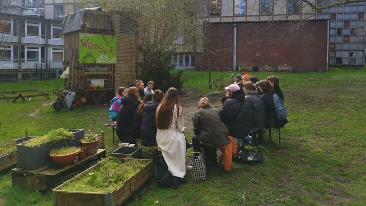 Eine Gruppe von ca. 15 Studis sitzen in einem Kreis auf Bierbänken am Wurzelwerk. Im vordergrund sieht man ein paar Hochhäuser mit Gewächshaus und den Geräteschuppen im Hintergrund. Auf einem Schild steht Wurzelwerk