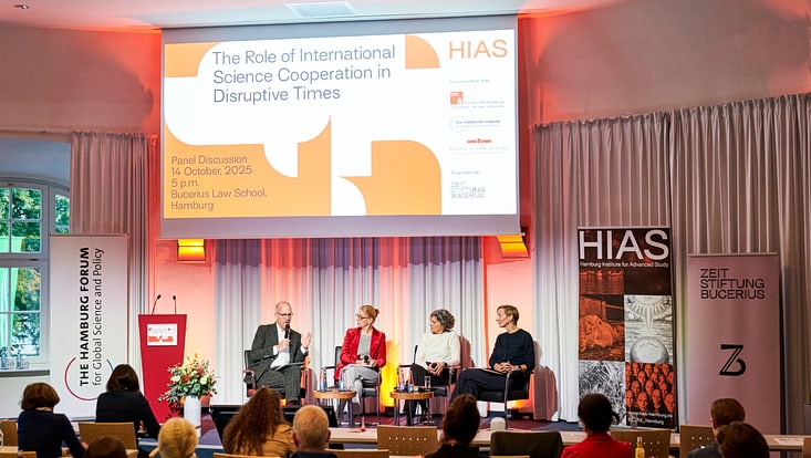Eine Podiumsdiskussion mit vier Personen findet in einem Saal statt. Auf der Bühne sitzen die Teilnehmenden vor einem großen Bildschirm, auf dem das Thema „The Role of International Science Cooperation in Disruptive Times“ und die Veranstalter „HIAS“